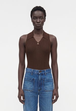 Γυναίκα με κοντά σγουρά μαλλιά που φοράει καφέ αμάνικη ριμπ μπλούζα με γιακά και ψηλόμεσο τζιν μπλε με μεγάλες μπροστινές τσέπες.