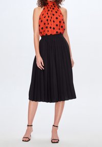 Top rosso con scollo all'americana e pois neri, abbinato a una gonna midi plissettata nera. L'outfit presenta una texture di tessuto uniforme e spalline sottili.