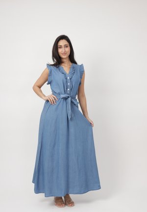 Kvinde stående med hånden på hoften iført blå maxi-kjole uden ærmer med knapper, flæsekrave, bundet bælte og sandaler mod ensfarvet baggrund.
