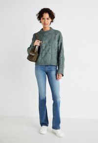 Maglione verde lavorato a maglia con motivo a treccia, abbinato a jeans svasati di un blu chiaro e sneakers bianche. Piccola borsa a tracolla verde oliva.