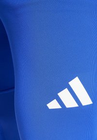 Blauwe sportlegging gemaakt van rekbaar materiaal, met een witte drie-strepen logo op het onderbeen. Glad oppervlak en aangesloten ontwerp.