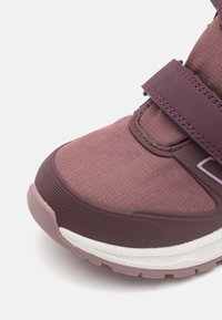 Fioletowe tekstylne sneakersy z teksturowanym wykończeniem, regulowanym paskiem, białą podeszwą oraz wzmocnionym przodem. Design podkreśla komfort i wsparcie.