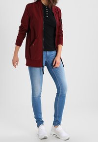 Burgundin värinen bomber-takki, jossa on etuvetoketju, sivutaskut ja ribbitaskut, yhdistetty mustan polo-paidan ja vaaleansinisten kapeiden farkkujen kanssa.