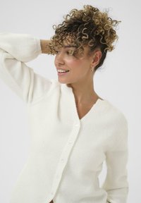 Frau mit lockigem Haar in einem weißen, geknöpften Cardigan, die lächelt und zur Seite blickt, mit einem Arm hinter ihrem Kopf erhoben.