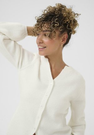 Femme aux cheveux bouclés portant un cardigan blanc à boutons, souriante et regardant sur le côté, un bras levé derrière sa tête.