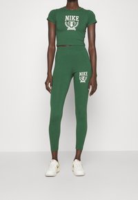 Žena ve zeleném zkráceném tričku Nike a legínách s bílými a žlutými teniskami Nike stojí na bílém pozadí.