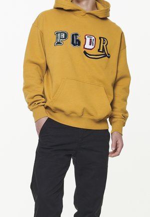 Persona con sudadera con capucha color mostaza y pantalones negros, la sudadera tiene letras de parches multicolores y un bolsillo frontal tipo canguro.