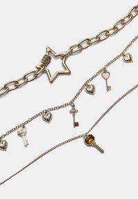 Trire ogrlice s zlatnim lancima. Jedna ima privjesak u obliku zvijezde, dok druge imaju privjeske u obliku srca i ključa, s različitim teksturama.