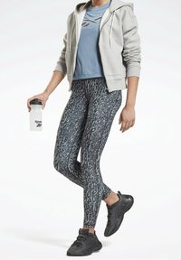 Atletisk outfit med en grå zip-up hoodie, blå t-shirt, mönstrade leggings i mörka färger och svarta sneakers. Håller en klar vattenflask.