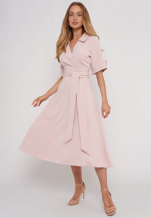 Kobieta ubrana w bladoróżową, midi sukienkę kopertową z rękawami do łokcia oraz paskiem w talii, zestawioną z beżowymi sandałami na wysokim obcasie z paskiem wokół kostki.