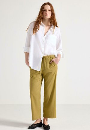 Camicia button-up bianca in lino, pantaloni verdi oliva con vita elastica e scarpe nere slip-on con design ritagliato. Outfit semplice e rilassato.