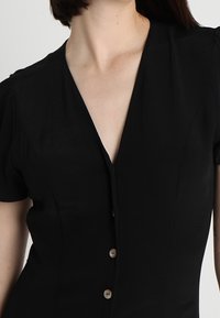 Černé šaty s krátkými rukávy, hlubokým výstřihem do V a pěti knoflíky vpředu. Hladká tkanina s přiléhavým střihem a jemnými detaily na ramenou.