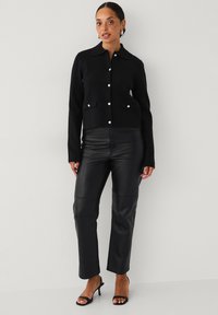 Zwarte gebreide jas met kraag en voorzakken, voorzien van witte knopen. Gecombineerd met zwarte leren broek en zwarte hakken.