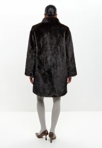 Mørk faux fur frakke med høj krave, lange ærmer og rundet kant, parret med grå ribbede strømpebukser og brune højhælede sko.