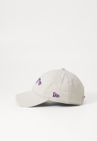Beige baseball cap i bomull med en buet skjerm, med lilla broderte tekst og logo på fronten og siden. Seks-panel design.