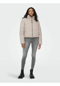 Giacca aviatore beige con colletto rialzato, chiusura frontale a zip e design trapuntato. Abbinata a jeans grigi e stivali neri alla caviglia.