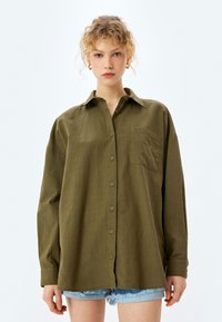 Camisa de botones verde oliva con manga larga, cuello y un bolsillo en el pecho; hecha de tejido texturizado. Llevada con pantalones cortos de mezclilla.