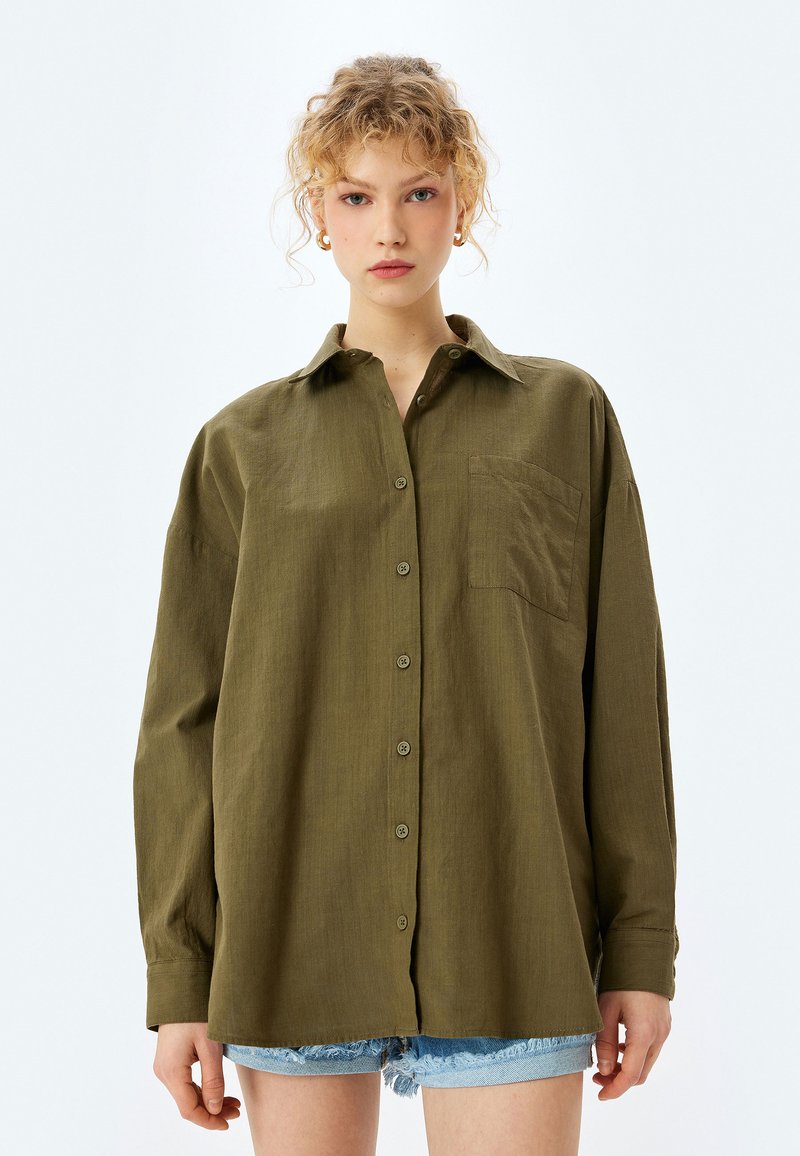 Camisa de botones verde oliva con manga larga, cuello y un bolsillo en el pecho; hecha de tejido texturizado. Llevada con pantalones cortos de mezclilla.