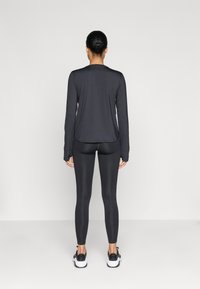 Tricou negru cu mâneci lungi, cu o croială lejeră, asortat cu colanți negri strâmți și adidași negri, pe un fundal neutru.