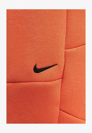 Tejido texturizado naranja con un logotipo de Nike negro bordado. Presenta un diseño suave y atlético adecuado para ropa deportiva.