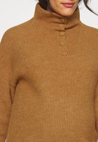 Femme portant un pull en maille couleur camel avec un col montant boutonné et un tissu texturé.