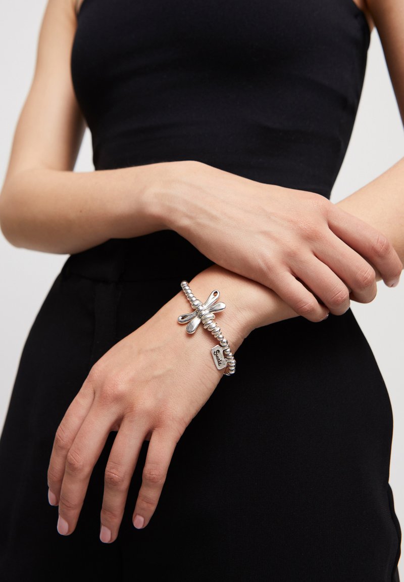 Bracelet en argent avec un design perlé et un pendentif libellule. Comprend une petite étiquette gravée du mot "Sister". Porté à la main sur un fond sombre.