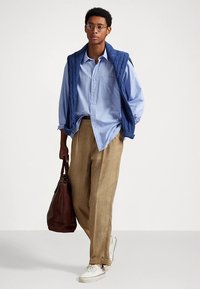 Ανοιχτό μπλε πουκάμισο με κουμπιά κάτω από μπλε καπιτονέ γιλέκο, μπεζ παντελόνι καρό, λευκά αθλητικά παπούτσια και καφέ τσάντα. Καθαρές γραμμές και χαλαρή εφαρμογή.