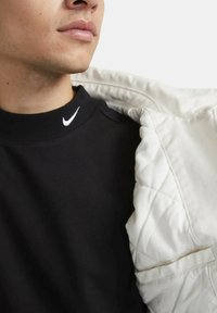 Sudadera negra con un logo blanco de Nike en el cuello, parcialmente cubierta por una chaqueta acolchada beige claro con un detalle de bolsillo.