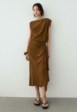 Frau trägt ein ärmelloses, braunes, drapiertes Midikleid mit seitlicher Bindung und schwarzen Plateau-Sandalen und steht vor einer schlichten weißen Wand.