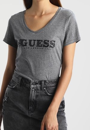Harmaa v-pääntie t-paita, jossa on musta koristeltu "GUESS" logo rinnassa. Kangas on pehmeä ja rento malli. Yhdistetty mustaan farkkuhameeseen.