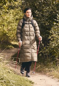 Gai olīvzaļš puffa mētelis ar kapuci, ar rāvējslēdzēja kabatām un quiltēta dizaina. Melni bikses, trekinga nūjas un izturīgas pārgājienu zābaki.