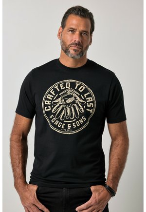 Mężczyzna z krótkimi włosami i brodą, noszący czarną koszulkę z beżową grafiką rękawic i tekstem "Crafted to Last Forge & Sons", stojący z rękami w kieszeniach.