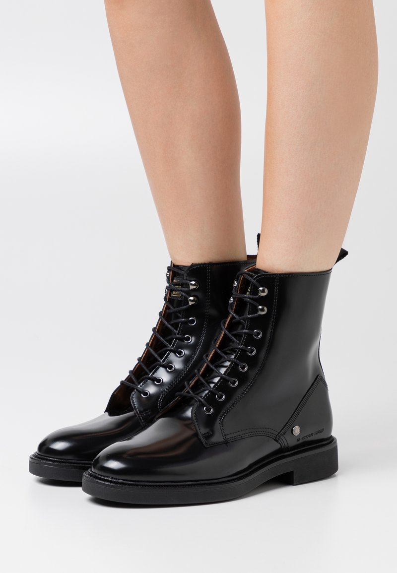 Bottines noires vernies à lacets portées sur des jambes nues sur un fond clair uni.