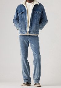 Homme portant une veste en velours côtelé bleu à doublure en polaire blanche, un pantalon en velours côtelé assorti, un pull crème et des baskets noires à lacets blancs.