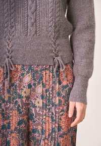 Suéter gris de punto con patrón de trenzas y lazos laterales, combinado con una falda floral plisada con colores cálidos y detalles de diseño intrincados.