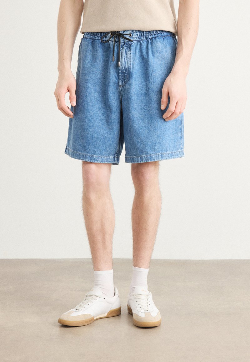 Shorts en jean bleu clair avec une taille élastique et un cordon de serrage, présentant une coupe décontractée et des ourlets retroussés. Des baskets blanches et des chaussettes de cheville sont portées.