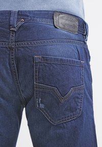 Jeans in denim blu scuro con una tasca posteriore caratterizzata da un motivo di cuciture a forma di V. Include una toppa in pelle con il marchio sulla cintura.