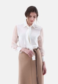 Blusa bianca con maniche trasparenti e un nastro annodato al colletto, abbinata a una gonna marrone con dettaglio della cerniera laterale.