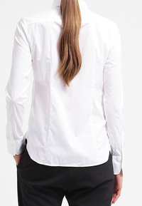 Camicia bianca a maniche lunghe, con bottoni, dal design aderente, con cucitura della spalla posteriore e orlo curvo, abbinata a pantaloni neri. Tessuto liscio, senza fantasie.