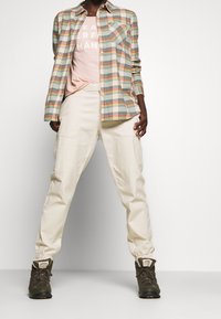 Person in cream-colored jogger pants, olive green hiking boots, peach t-shirt partially covered by open plaid shirt in soft earth tones.