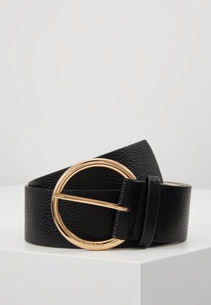 Czarny teksturowany skórzany pasek zwinięty na białej powierzchni z dużą, okrągłą, polerowaną złotą klamrą.