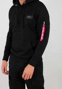Uomo che indossa un hoody nero con il logo Alpha Industries e un'etichetta rossa "Rimuovere Prima del Volo" sulla cerniera della tasca sinistra della manica.