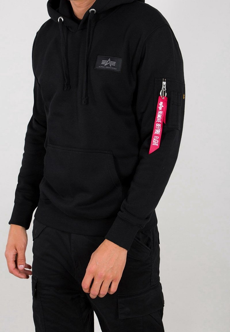 Uomo che indossa un hoody nero con il logo Alpha Industries e un'etichetta rossa "Rimuovere Prima del Volo" sulla cerniera della tasca sinistra della manica.