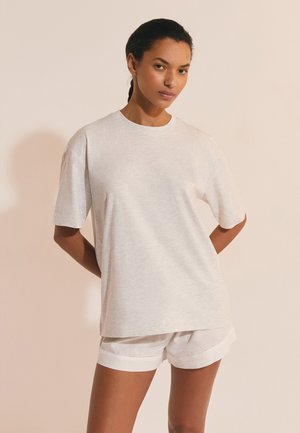Femme aux cheveux tressés portant un T-shirt ample gris clair et un short assorti de couleur claire, sur un fond beige uni.