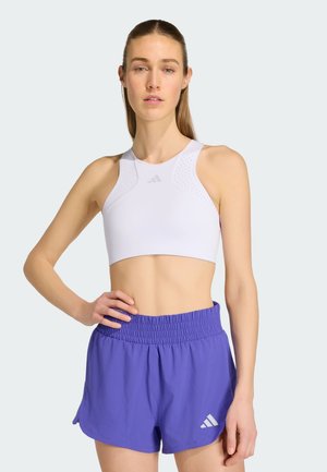 Frau trägt ein weißes ärmelloses Sport-Crop-Top und blaue sportliche Shorts, steht mit einer Hand in der Hüfte vor einem einfarbigen Hintergrund.