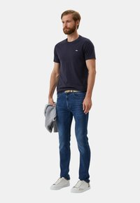 T-shirt navy, jeans di denim blu slim-fit, felpa grigia drappeggiata su un braccio, sneakers bianche con dettagli sottili, in posa eretta contro uno sfondo neutro.