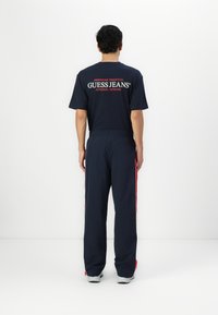 Tamsiai mėlyna marškinė su balta ir raudona "GUESS JEANS" užrašu ant nugaros, derinami su tamsiai mėlynomis kelnėmis su raudonomis šoninėmis juostomis. Minkšta medžiaga.