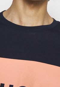 Námořnická modrá tričko s kulatým výstřihem, s růžovým grafickým prvkem přes hruď a tučným černým textem. Měkká bavlněná tkanina, volný střih.