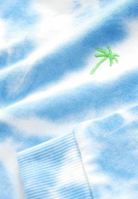 Blå tie-dye bomullströja med vita molnmönster. Har en liten grön palmträdsembroidery och ribbade ärmslut. Mjuk textur.