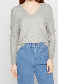 Pull gris à col en V en maille texturée, associé à un jean taille haute bleu clair avec bouton et poches classiques.